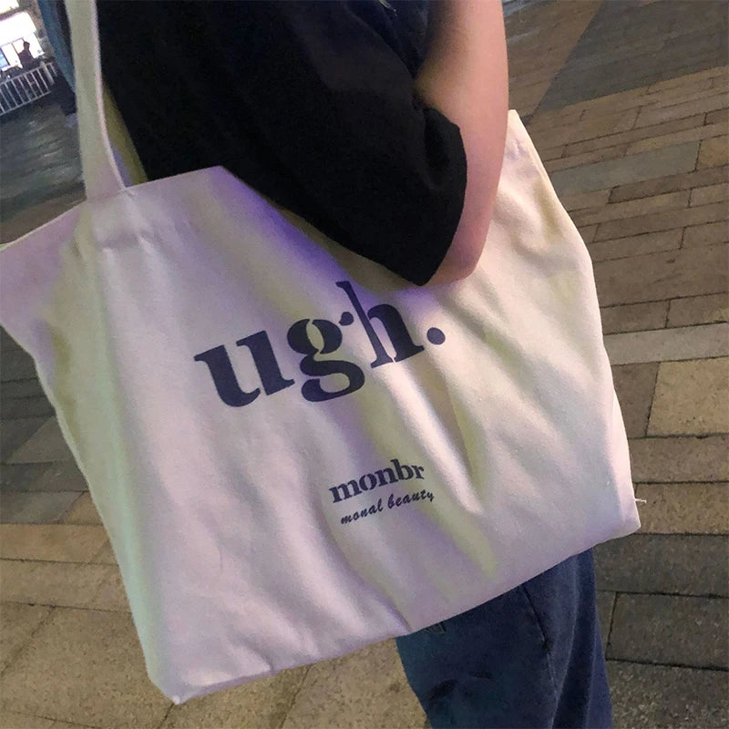 Tote bag with 'ugh.' text held by a person on a pavement.