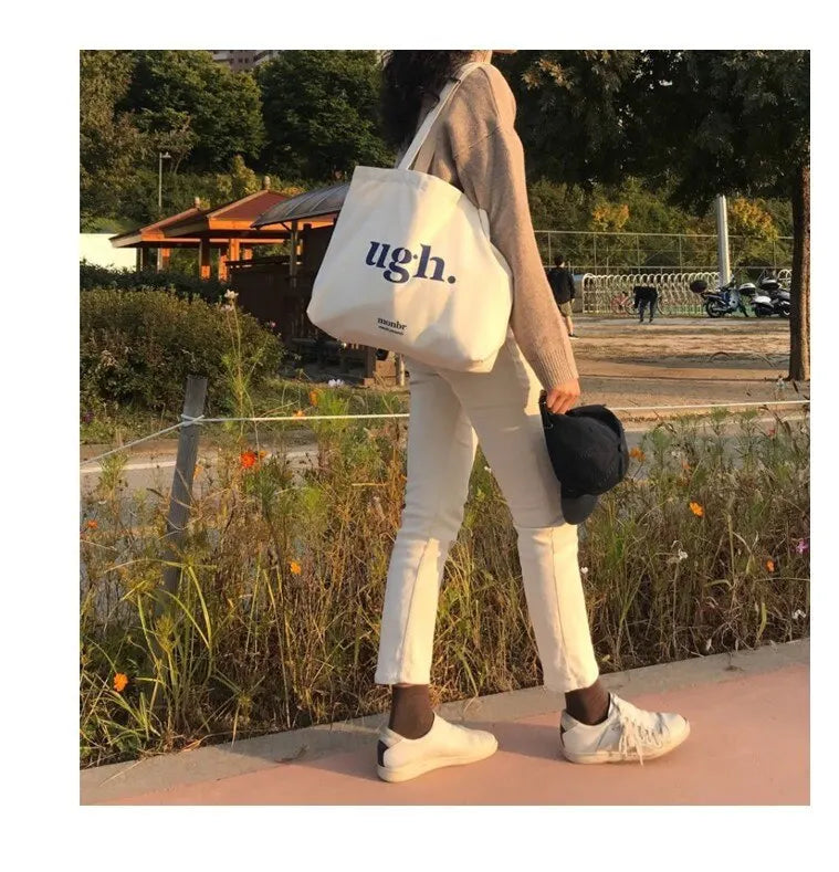 Person walking outdoors with a white tote bag labeled 'ugh.'