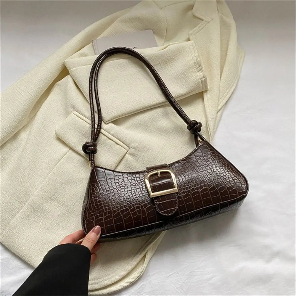 Brown crocodile-patterned handbag held by a hand against a beige coat on a light gray background