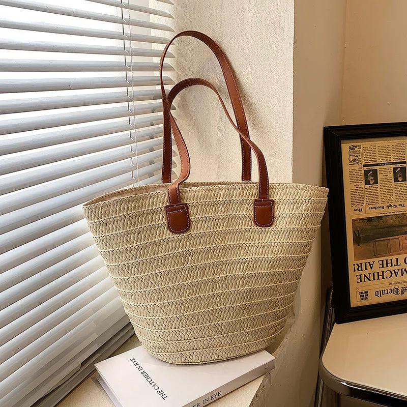 Woven beige tote bag with brown handles on a surface near a window with blinds.