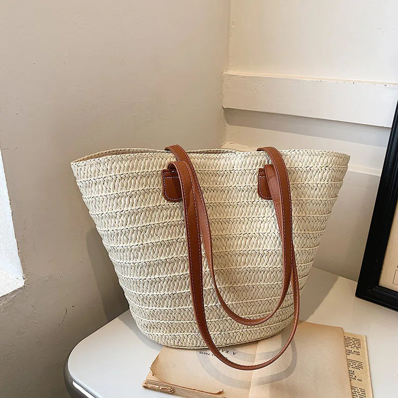 Woven straw tote bag with brown leather straps on a white surface.