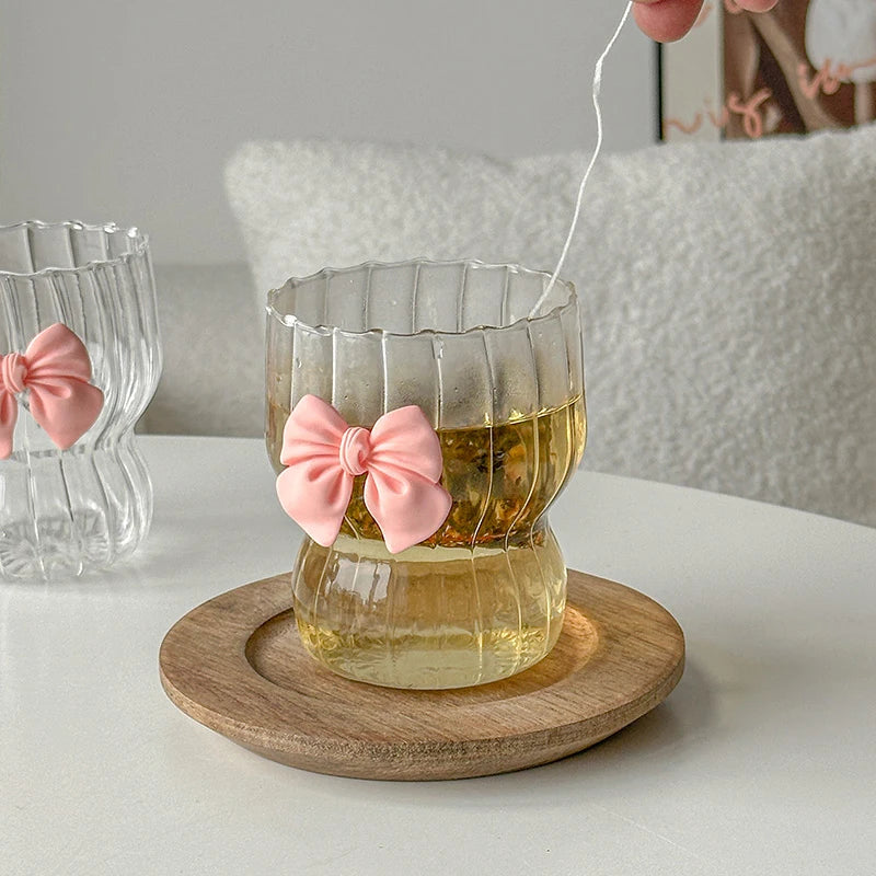 Clear glass with pink bow and tea bag on a wooden coaster, with another glass in the background.