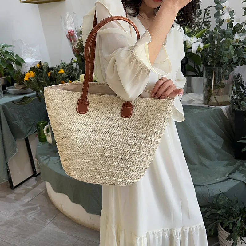 Person holding a woven beige tote bag with brown handles in an indoor setting with plants.