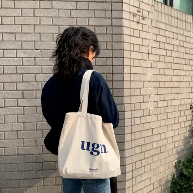 Person carrying a tote bag with 'ugh' text against a brick wall