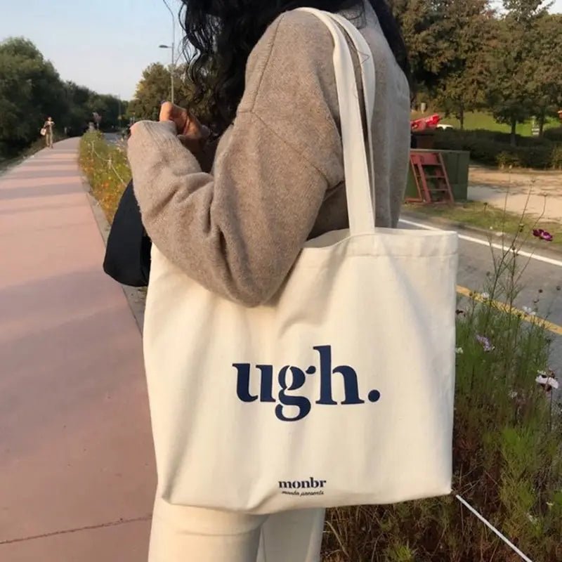 Person holding a tote bag with 'ugh.' text outdoors