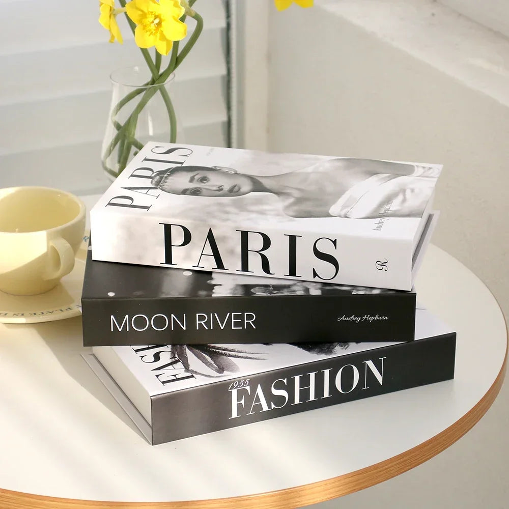 Stack of decorative books titled 'Paris', 'Moon River', and 'Fashion' on a table with a cup and yellow flowers.