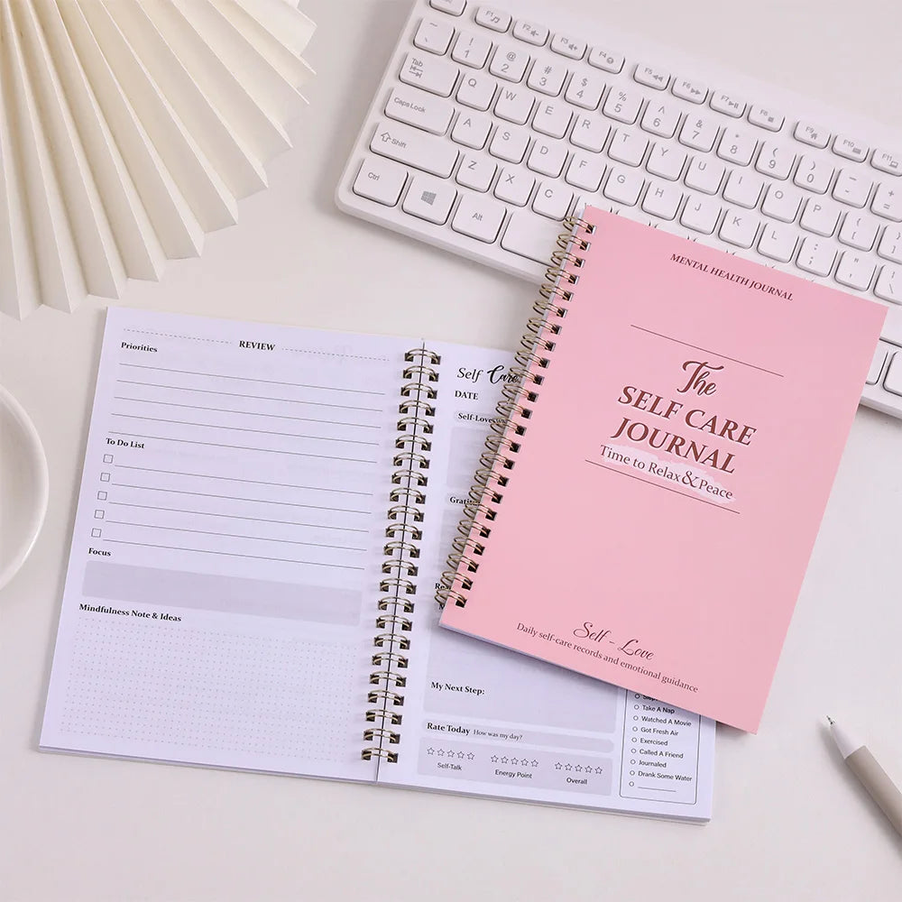 Pink self-care journal open on a desk with a keyboard and pen