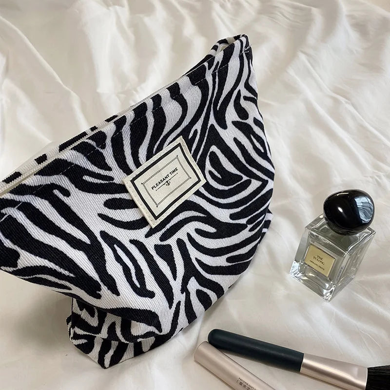 Zebra print pouch with a label, next to a perfume bottle and makeup brush on a white surface.