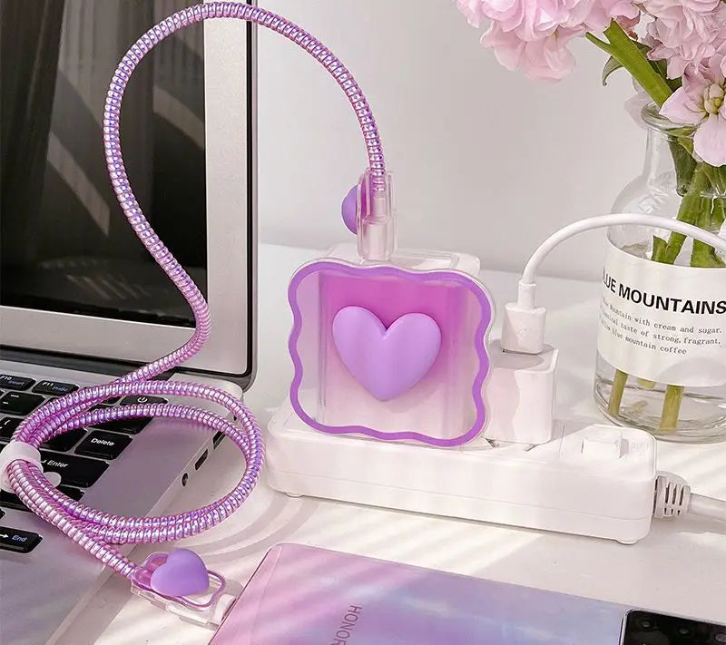 Purple heart-shaped charger head protective sleeve with flexible cable on a desk next to a laptop and flowers.
