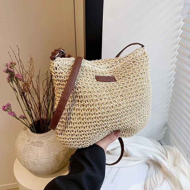 Woven handbag with brown handle held by a person, with a vase of flowers and white curtain in the background.