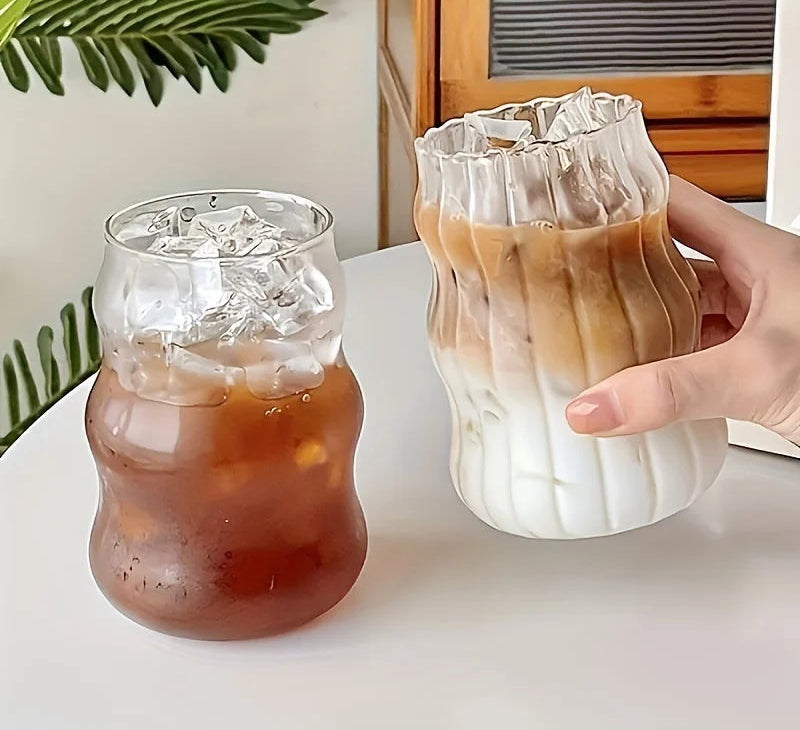 Two glass tumblers with iced coffee and a hand holding one of them on a white surface.